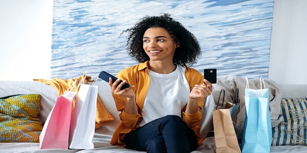 Happy shopper sitting on a sofa with multiple shopping bags, using her smartphone and bank card—representing loyal customers who become brand ambassadors through rewarding loyalty programs.